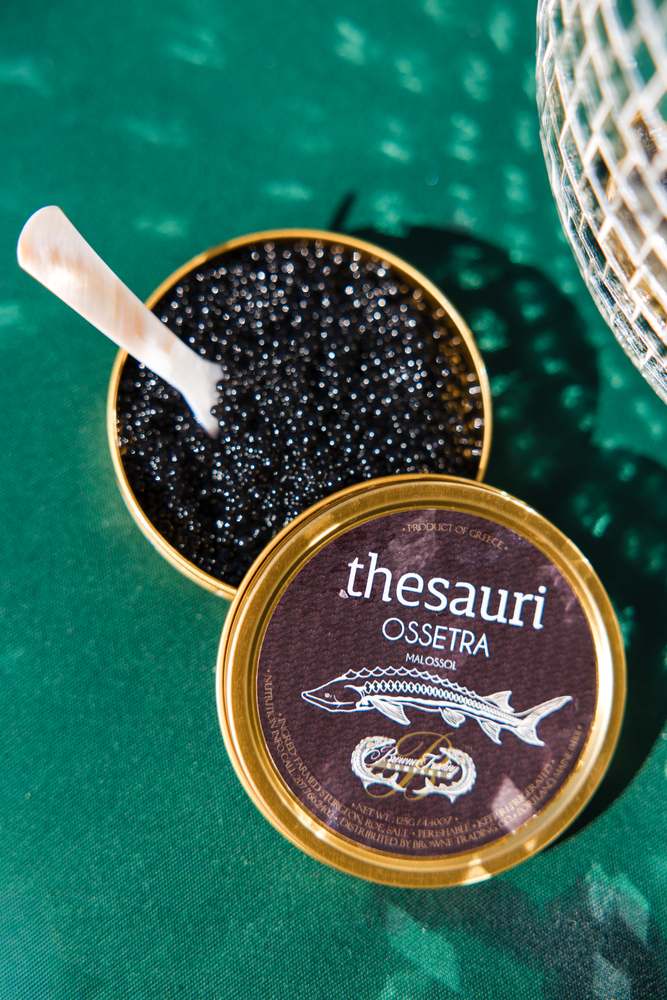 An open gold tin of Thesauri Ossetra caviar with a mother-of-pearl spoon, on green.