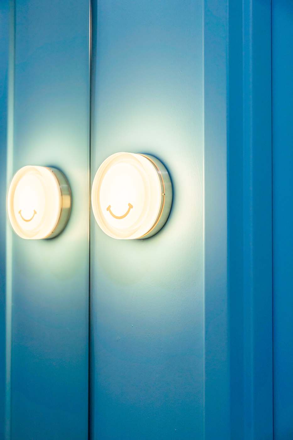 Two round sconces with a smile embossed on the glass, lit against the blue panelled wall at Squeeze.