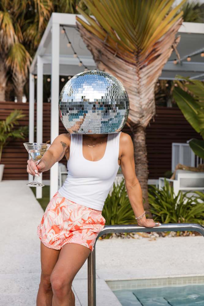 Poolside in coral-print shorts and a white tank, mirror-ball head, martini raised.