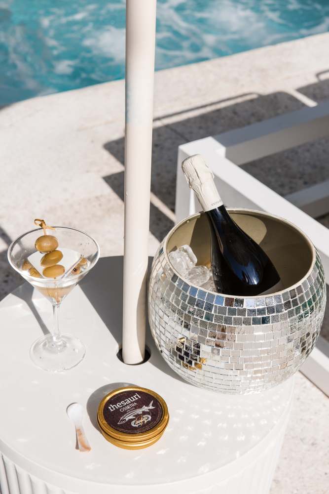 A martini with olives, a mirror-ball ice bucket holding prosecco, and a gold tin of Thesauri Ossetra caviar on a white table.