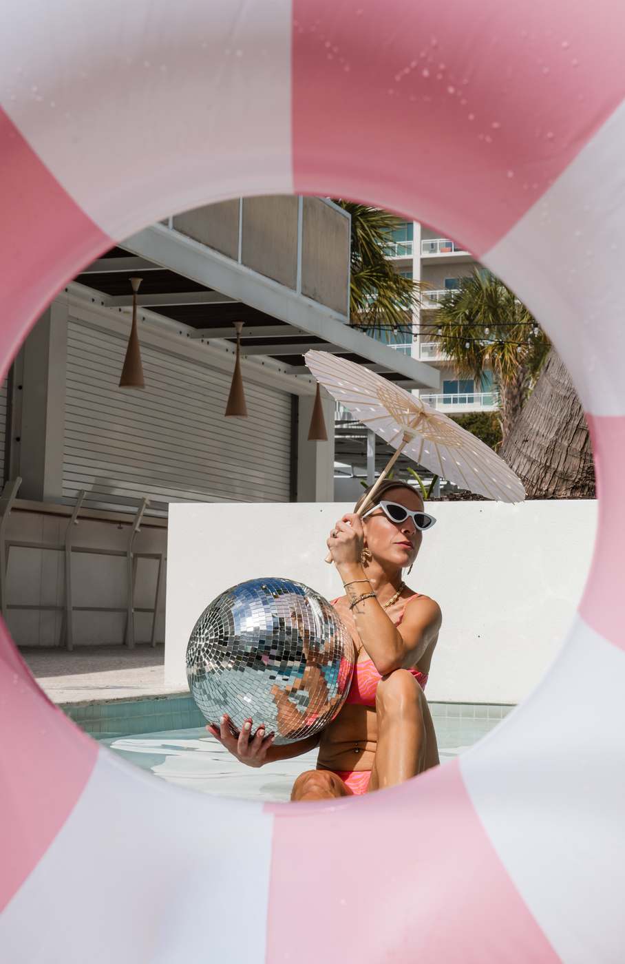 Framed through a pink and white ring float: sitting at the water's edge holding the mirror ball, parasol open behind.