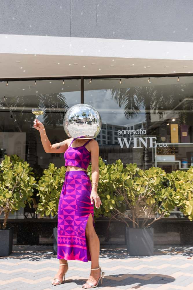 Outside Sarasota Wine Co. on Palm Avenue — the full storefront logo in the window — mirror-ball head, coupe raised.