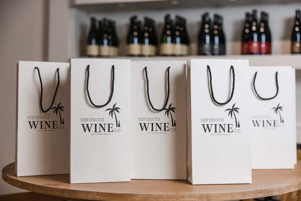 A row of white Sarasota Wine Co. gift bags with black rope handles on a round wooden table.