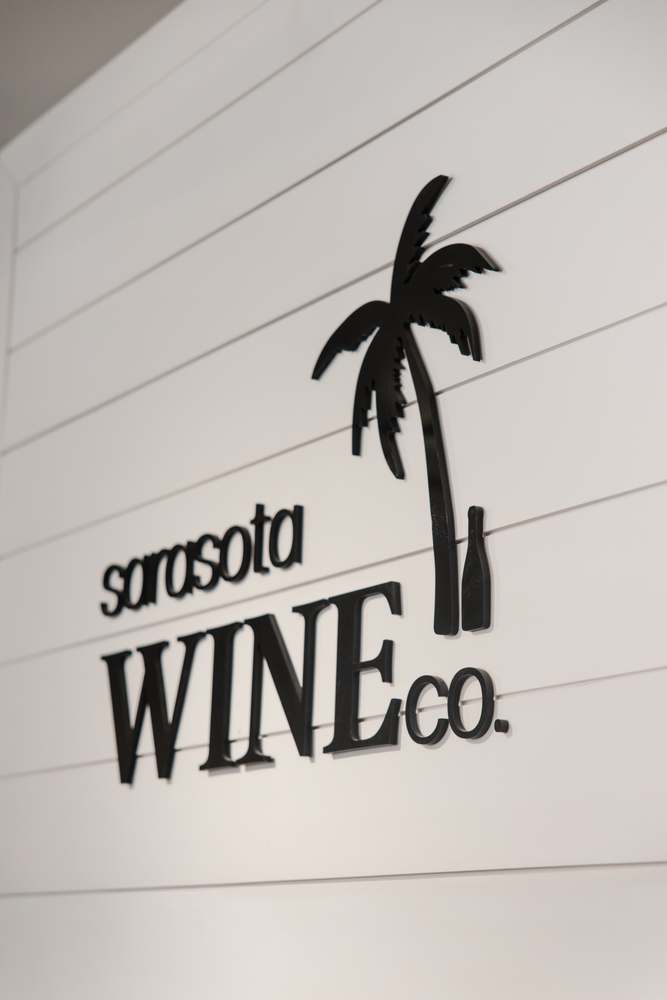 The Sarasota Wine Co. logo — palm tree and bottle — mounted on a white shiplap wall.
