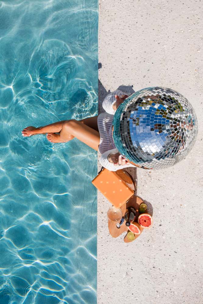 Overhead: the mirror-ball head sitting at the pool's edge, feet in the water, the tile line splitting the frame.