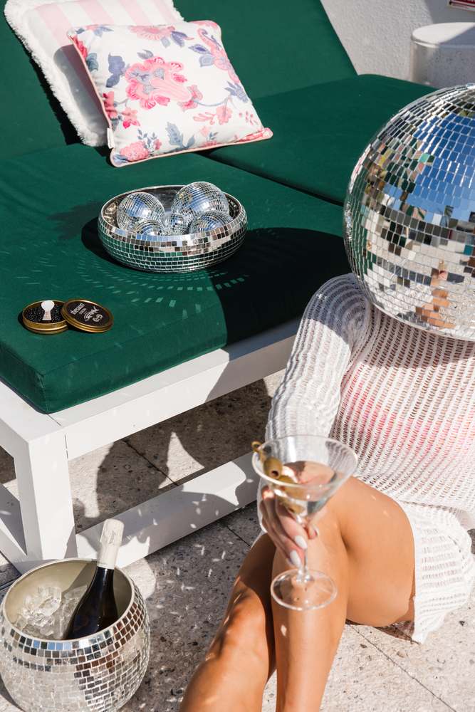 A green poolside lounger with caviar tins and a bowl of small mirror balls, mirror-ball head and a martini in hand.